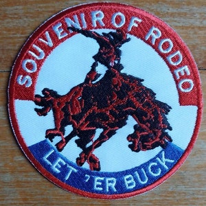Op de afbeelding: Een geborduurde patch in rood, wit en blauw met een cowboy die een wild paard berijdt. De tekst "Souvenir of Rodeo" staat in een rode cirkel rond de afbeelding. De tekst "Letter Buck" staat in een blauwe cirkel onder de afbeelding.