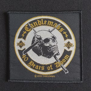 Könnte beinhalten: Schwarzer quadratischer Aufnäher mit einem kreisförmigen Design, das einen Totenkopf mit gekreuzten Knochen zeigt. Der Text "Candlemass" wölbt sich über dem Totenkopf, darunter "40 Years of Doom". Die Daten "1983" und "2023" sind ebenfalls vorhanden. Der Aufnäher hat goldene und weiße Akzente.
