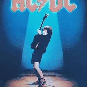 May include: Black t-shirt featuring the band AC/DC's logo in orange and a guitarist playing. The text "LET THERE BE ROCK" is below the image. The background is blue with a spotlight effect.