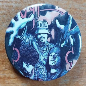May include: Circular button featuring a graphic of three men in shades of dark blue and pink. One man is wearing a bucket hat with the word "KILL". The button has a vintage, slightly distressed appearance.
