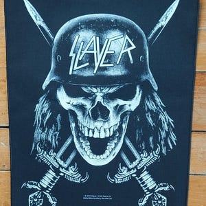 May include: Black patch featuring a skull wearing a helmet with the word "SLAYER" in white. Two crossed swords are behind the skull. The patch has a white and black design.