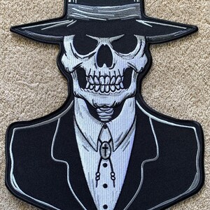 May include: A black and white embroidered patch of a skull wearing a black fedora and a suit. The skull has a white tie with a silver cross pendant.