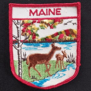 Könnte beinhalten: Ein weißer gestickter Aufnäher mit rotem Rand. Der Aufnäher zeigt eine Landschaft mit einem See, Bäumen und einem Reh mit einem Kitz. Der Text "MAINE" ist oben auf dem Aufnäher in Rot gestickt.