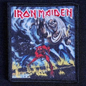 May include: A rectangular patch featuring the band name "IRON MAIDEN" in red letters. The patch depicts a skeletal figure with long white hair, reaching out, and a red devil-like creature. The background is a dark blue with lightning.