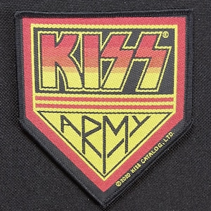 Pode incluir: Um patch preto e vermelho com o logotipo da banda KISS em um gradiente de amarelo a vermelho. Abaixo do logotipo está a palavra "ARMY" em amarelo, em um fundo vermelho e preto. O patch é contornado em preto.