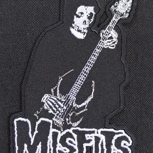 Pode incluir: Patch bordado em preto e branco com uma figura esquelética tocando baixo. A figura está usando uma capa com capuz. A palavra "MISFITS" é bordada em branco, com contorno preto, abaixo da figura.