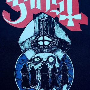 May include: Black graphic featuring the band name "Ghost" in red gothic lettering above a detailed illustration. The illustration includes a skull, a papal mitre, and figures in black robes, with a blue and white colour scheme.