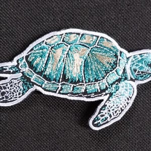 Pode incluir: Um remendo bordado de tartaruga marinha em azul turquesa e branco. A tartaruga está virada para a direita e tem uma carapaça e nadadeiras detalhadas.