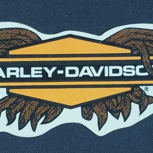May include: A die-cut sticker featuring the Harley-Davidson logo in white text on a black rectangular bar, centered over a yellow hexagon. Large, detailed brown wings with visible feather textures extend from both sides of the central emblem.