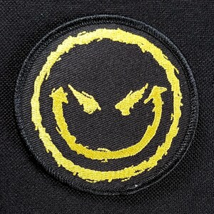 May include: A black fabric patch with a yellow embroidered smiley face with an angry expression. The smiley face is outlined in black.