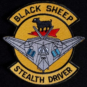 Może przedstawiać: Haftowana naszywka z żółtą obwódką i czarnym tłem. Naszywka zawiera tekst "BLACK SHEEP" i "STEALTH DRIVER". Czarna owca znajduje się nad stylizowanym projektem samolotu.