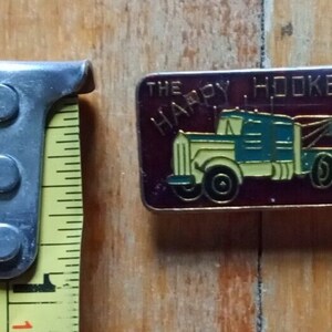 May include: A red, white, and blue enamel pin with a cartoon image of a yellow and blue semi-truck with a tow truck attached. The text "THE HAPPY HOOKER" is printed in white on a red background.