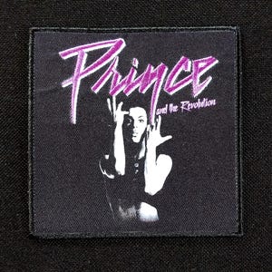 May include: A black square patch featuring a stylized image of Prince with his hands raised. The name "Prince" is written in purple script above the image, with "and the Revolution" below.
