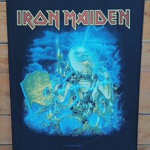 May include: Black rectangular patch featuring the band name "IRON MAIDEN" in gold lettering. The image depicts a skeletal figure with blue energy effects, standing near a tombstone. The background is dark blue with lightning.