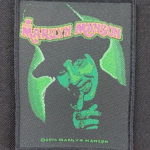 May include: A rectangular black patch featuring the text "MARILYN MANSON" in pink and orange, above a green and black graphic of a face and hand. The text "©2016 MARILYN MANSON" is at the bottom.