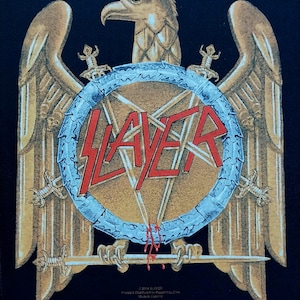 Op de afbeelding: Zwarte afbeelding met een gouden adelaar met uitgespreide vleugels, een schild met de bandnaam "SLAYER" in rood en gekruiste zwaarden. Het ontwerp bevat tekst onderaan die de band en de herkomst aangeeft.