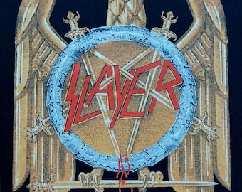 以下が含まれることがあります： 黒いグラフィック。金色のワシが翼を広げ、赤い「SLAYER」のバンド名が入った盾と交差した剣が描かれています。デザインには、バンドと原産地を示すテキストが下部に含まれています。