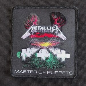 May include: A square embroidered patch with a black background featuring the Metallica logo in white above a graphic of white crosses in a green field under a red and yellow sky, with the text MASTER OF PUPPETS printed in white at the bottom.