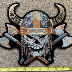May include: A black embroidered patch with a skull wearing a horned helmet and crossed axes. The skull is white with black eyes and a black beard. The helmet is gold and silver with a black and white braided design. The axes are silver with black handles.
