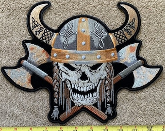 May include: A black embroidered patch with a skull wearing a horned helmet and crossed axes. The skull is white with black eyes and a black beard. The helmet is gold and silver with a black and white braided design. The axes are silver with black handles.