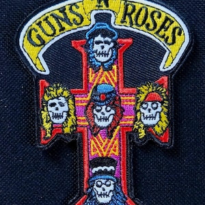 May include: Embroidered patch featuring the Guns N' Roses logo. The design includes a cross shape with skull faces, and the band's name in yellow lettering on a black background. The patch has a red, pink, and yellow color scheme.