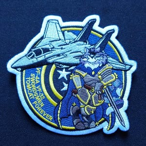 Puede incluir: Parche bordado con un gato de dibujos animados con armadura sosteniendo una espada, con un avión de combate volando por encima. El parche tiene un fondo azul con un círculo amarillo y el texto "VF-44A VF-32 SWORDSMEN TOMCAT FOREVER".
