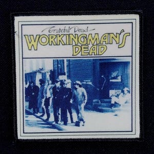 Può includere: Una toppa rettangolare con bordo bianco e sfondo nero. La toppa presenta il testo "Grateful Dead WORKINGMAN'S DEAD" in giallo e un'immagine blu e bianca di persone davanti a un edificio.