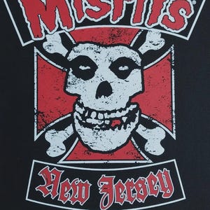 Może przedstawiać: Czarny grafika z czerwonym napisem "MISFITS" nad białą czaszką z skrzyżowanymi kośćmi na czerwonym tle. Poniżej tekst "New Jersey" i "EST. 1977" wyświetlony w kolorze czerwonym i białym, klasyczny design punk rock.