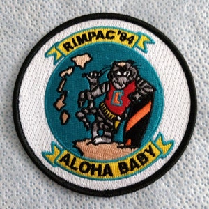 May include: A white embroidered patch with a black border. The patch features a cartoon tiger wearing a red shirt with a white letter 'E' on it. The tiger is holding a surfboard and standing on a beach with the text 'Aloha Baby' and 'RIMPAC '84' around it.