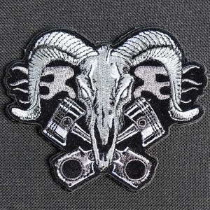 May include: A silver embroidered patch of a ram skull with crossed pistons. The ram skull is detailed with horns and a prominent nose. The pistons are stylized and have a black background.