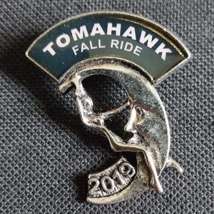 May include: A silver metal pin with a black and white design. The pin features a crescent moon with a figure climbing up it. The text "Tomahawk Fall Ride 2019" is on the top of the pin.
