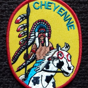 Puede incluir: Un parche ovalado amarillo bordado con un borde rojo. El parche presenta a un hombre nativo americano montando un caballo blanco con manchas negras. El hombre lleva un tocado azul y blanco y sostiene una lanza. El texto "CHEYENNE" está bordado en azul en el parche.