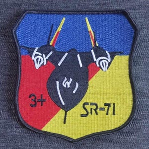 Puede incluir: Parche bordado con un diseño de escudo que presenta una silueta de avión negra sobre un fondo azul, rojo y amarillo. El parche incluye el texto "3+" y "SR-71" en blanco.