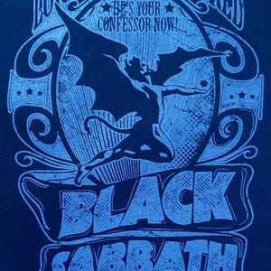 May include: A navy blue graphic tee featuring a stylized image of a winged figure and the words "LORD OF THIS WORLD" and "BLACK SABBATH" in light blue. The text "HE'S YOUR CONFESSOR NOW!" is also present.