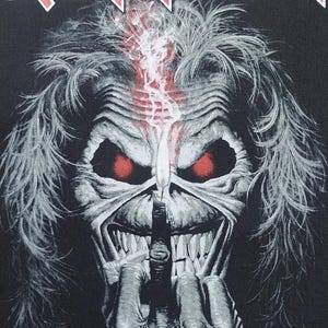 May include: Black graphic featuring the Iron Maiden band logo and a detailed illustration of their mascot, Eddie. Eddie has red eyes, a skeletal face, and a finger to his lips. The image has a dark, ominous aesthetic.