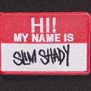 Pode incluir: Um patch retangular com a parte superior vermelha e a inferior branca. O texto "HI! MY NAME IS SLIM SHADY" é bordado em fio branco e preto. O patch tem uma borda vermelha e está sobre um fundo preto.