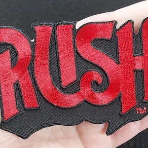Pode incluir: Etiqueta bordada vermelha com a palavra "RUSH" em uma fonte estilizada em um fundo preto.