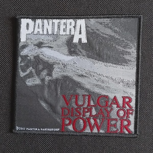 May include: Square patch featuring the band name "PANTERA" in white block letters above the phrase "VULGAR DISPLAY OF POWER" in red. The patch has a grayscale image background and the text "©2012 PANTERA PARTNERSHIP".