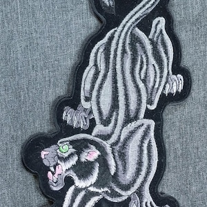 May include: Embroidered patch featuring a stylized panther design. The panther is depicted in shades of gray with black outlines, showcasing a striped pattern. The panther has green eyes and pink accents on its mouth and ears. The patch is on a gray background.
