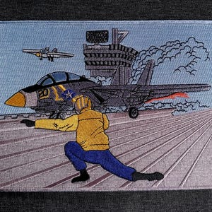 May include: Embroidered patch featuring a fighter jet on an aircraft carrier deck. The patch depicts a pilot in a yellow jacket and blue pants, directing the jet. A second jet is in the sky. The background includes a control tower and clouds.