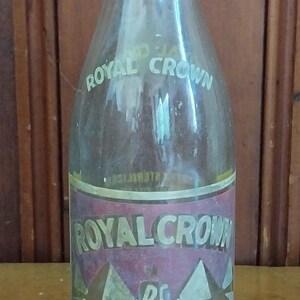 May include: Vintage Royal Crown Cola bottle. The clear glass bottle features a label with the words "Royal Crown" and "RC Cola". The label has a red and white design. The bottle contains 12 fluid ounces.
