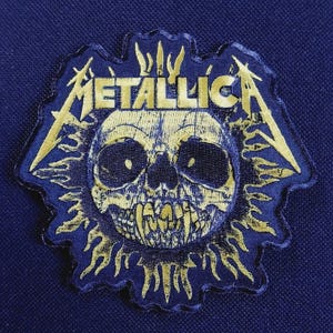 May include: A dark blue embroidered patch featuring a yellow skull graphic with sharp teeth and the word METALLICA in a stylized font. The design is surrounded by flame-like shapes, creating a jagged, irregular border on a dark blue fabric background.