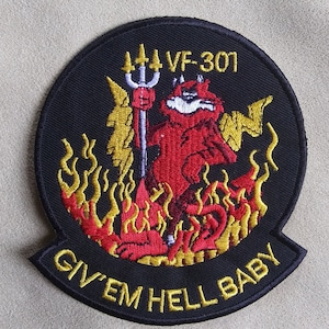 May include: A black embroidered patch with a red devil holding a trident, surrounded by yellow flames. The text "VF-301" and "Give 'em Hell Baby" are embroidered in yellow.