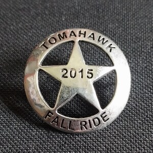 May include: A silver metal pin with a five-pointed star in the center. The star has the year "2015" inscribed inside. The words "Tomahawk Fall Ride" are inscribed around the star.