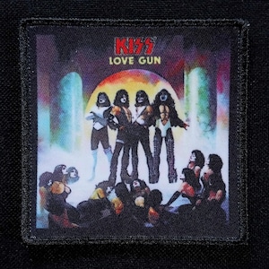 Pode incluir: Patch quadrado com a banda KISS e o texto "LOVE GUN". A imagem mostra os membros da banda com sua maquiagem e figurinos icônicos, com um fundo colorido. O patch é contornado com costura preta.