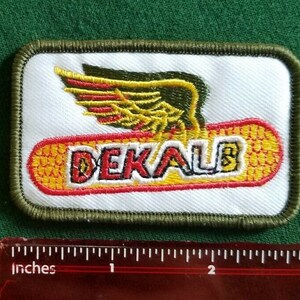 Peut inclure: Écusson rectangulaire à fond blanc, avec un motif ailé rouge et jaune au-dessus du mot "DEKALB" en rouge et jaune. L'écusson a une bordure verte et mesure environ 7.6 cm de long.