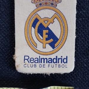 May include: A white patch with a gold and blue crest of Real Madrid Club de Futbol. The crest features a crown, a football, and the letter F.
