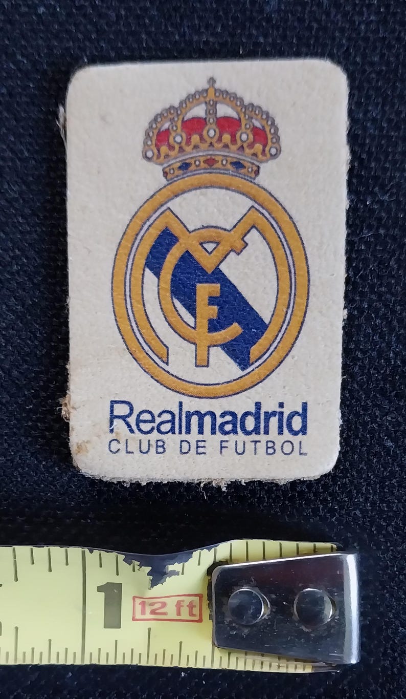 May include: A white patch with a gold and blue crest of Real Madrid Club de Futbol. The crest features a crown, a football, and the letter F.