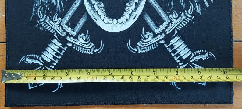 May include: Black rectangular patch with a white graphic of a skull, crossed swords, and bird talons. The patch is approximately 7 inches wide. A yellow measuring tape is placed across the bottom of the patch.