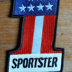 Puede incluir: Un parche bordado con forma de número uno, con los colores rojo, blanco y azul y la palabra "SPORTSTER" en letras negras sobre fondo blanco.